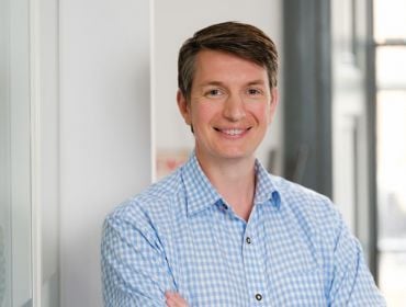 A man wearing a blue and white check shirt with his arms folded smiling at the camera. He is standing in a brightly lit glass office building.