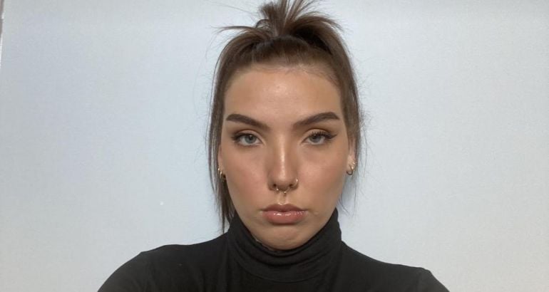 A woman wearing a dark turtleneck with her hair tied up looks into the camera in front of a grey wall. She is Mallaidh Downes, a cloud engineering consultant at Deloitte.