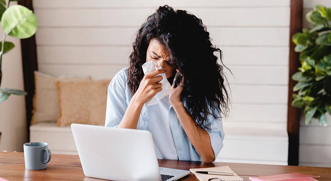 A young woman attempts to work from home while sick, she is on the phone and the laptop.