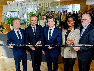 Multiple men and women wearing suits, cutting a ribbon inside a Proofpoint office.