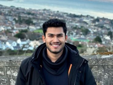 A young man wearing a black coat stands in front of a wall outside. In the background is a sprawling landscape view.