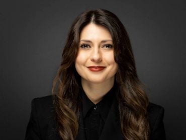 A headshot of a woman with long brown hair smiling at the camera against a charcoal background. She is Smarttech247 CEO Raluca Saceanu.