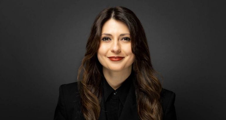 A headshot of a woman with long brown hair smiling at the camera against a charcoal background. She is Smarttech247 CEO Raluca Saceanu.