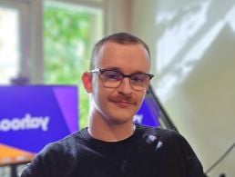 Yahoo's Nate McCoy stands against a white wall with a geometric pattern smiling at the camera. He is wearing an orange t-shirt.