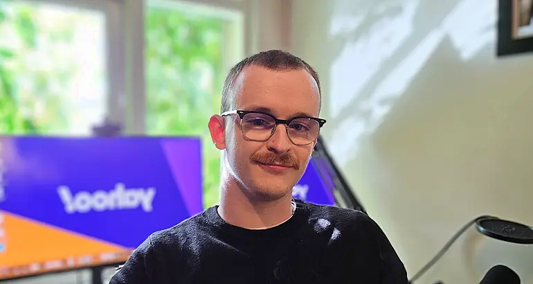 A man with a moustache wearing glasses and a dark T-shirt smiles at the camera while sitting in a home office, with a computer monitor placed behind him.