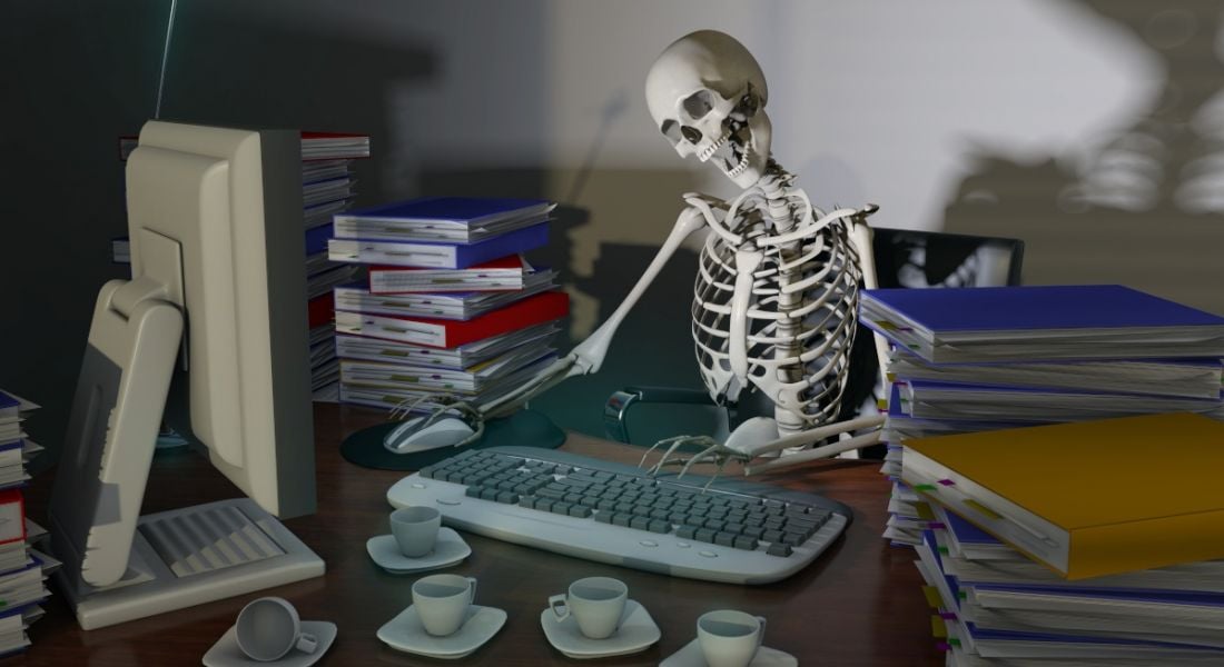 A skeleton works at a desk, representing the danger of overworking to physical, emotional and mental health.