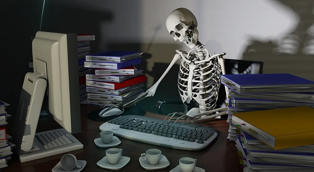 A skeleton works at a desk, representing the danger of overworking to physical, emotional and mental health.