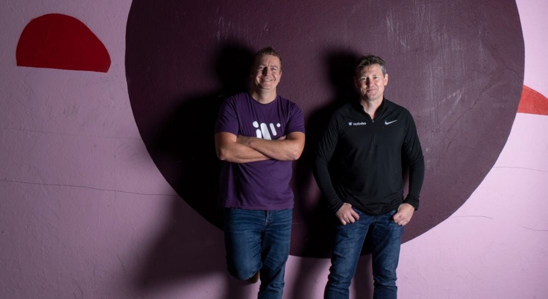 Two men stand up against a concreate wall painted pink with a different shapes on it. They are the co-founders of Workvivo.