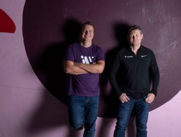 Two men stand up against a concreate wall painted pink with a different shapes on it. They are the co-founders of Workvivo.