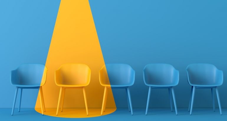 A yellow light shines on a blue room full of blue chairs, showing one person has been chosen for a job.