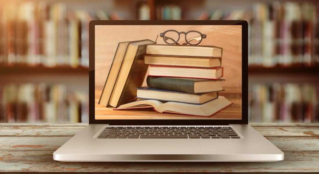 Books and glasses are displayed on a laptop, which is placed on a table in front of a bookshelf, indicating digital literacy.