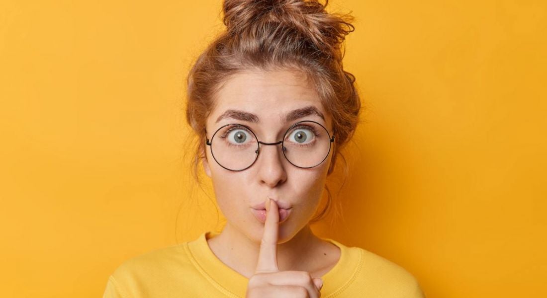 Woman in glasses, with a top bun, in yellow, is shushing, indicating what not to say.