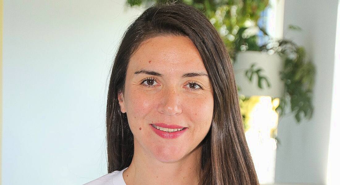 A woman with long brown hair smiles at the camera in front of a white wall and a large plant. She is Maria Duni, the country manager for the Nordics at Personio.