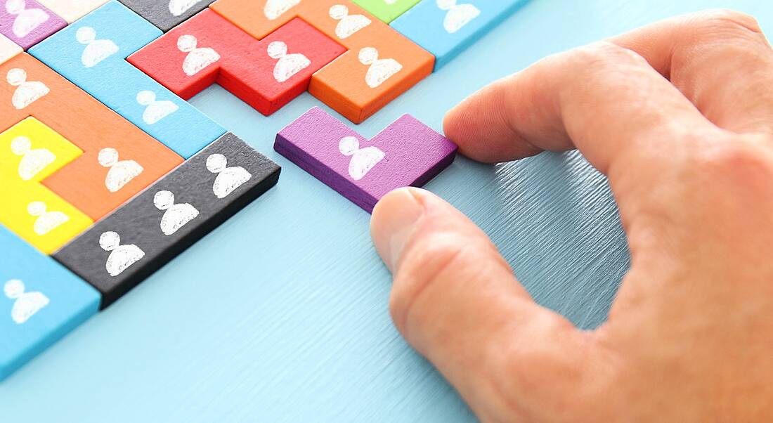 A person's hand holding a purple puzzle piece against a nearly completed multi-coloured jigsaw with figures painted on each piece in white. The background is blue.