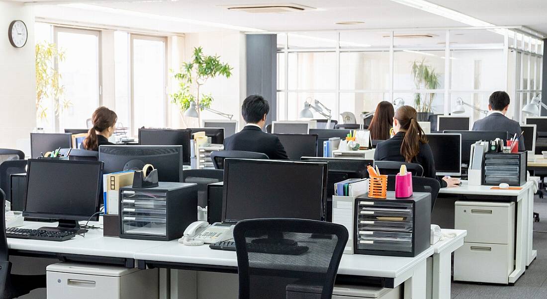 Employees in an office sitting at desks with black ergonomic chairs and computer monitors.