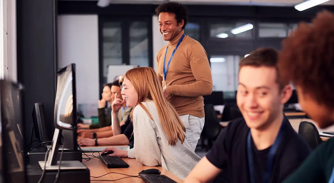 People learning tech skills in an office environment sitting at computers with a teacher standing behind them providing direction.