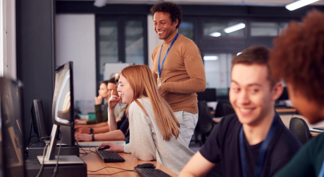 People learning tech skills in an office environment sitting at computers with a teacher standing behind them providing direction.