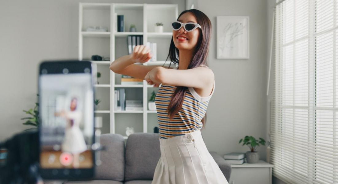 Young woman filming a TikTok video in a living room area. She is wearing sunglasses and moving energetically to a camera which is placed on a tripod.