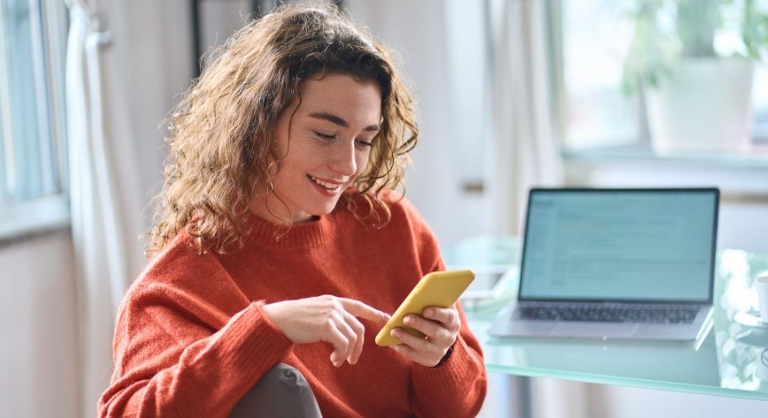 Woman using her smartphone while working. There is a laptop at her desk but she appears preoccupied by the mobile phone. She is smiling and happy.