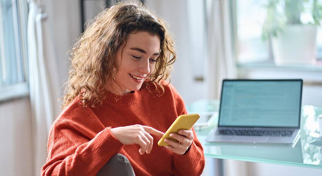 Woman using her smartphone while working. There is a laptop at her desk but she appears preoccupied by the mobile phone. She is smiling and happy.