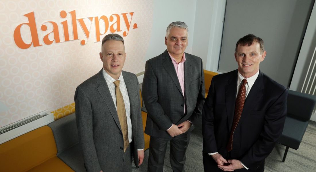 Three people standing in front of a wall with DailyPay logo on it in orange print.