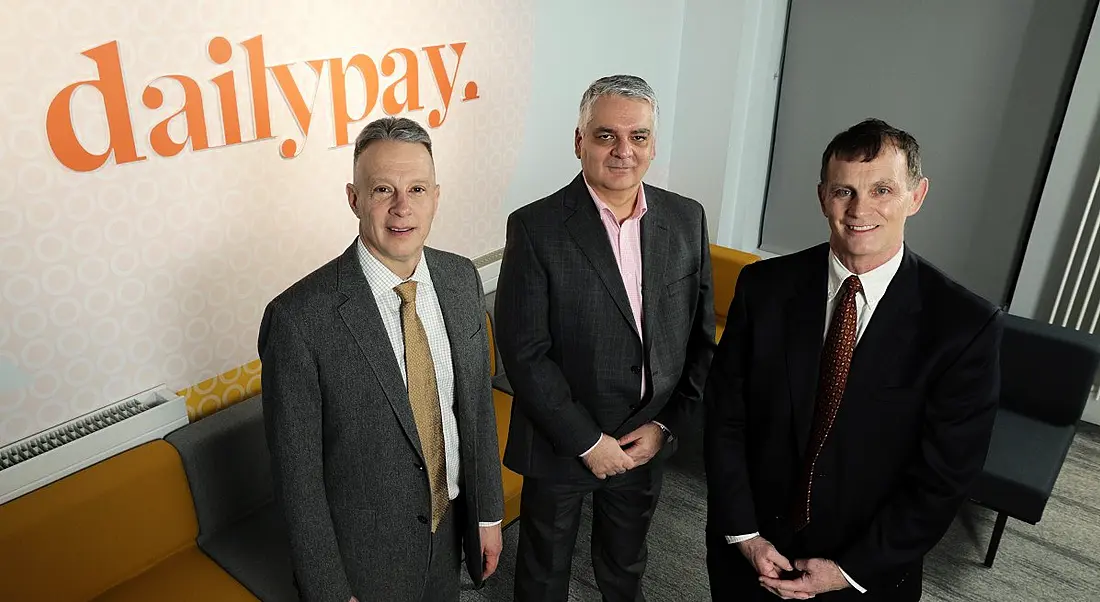 Three people standing in front of a wall with DailyPay logo on it in orange print.