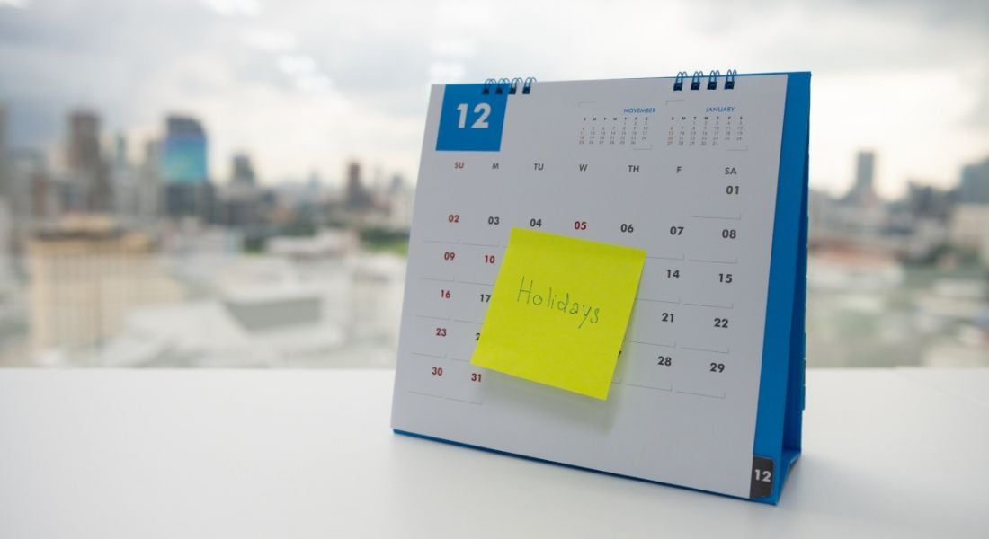 A calendar with a sticky note sits next to a window where a city skyline can be seen. The word 'Holiday' is written on the sticky note.
