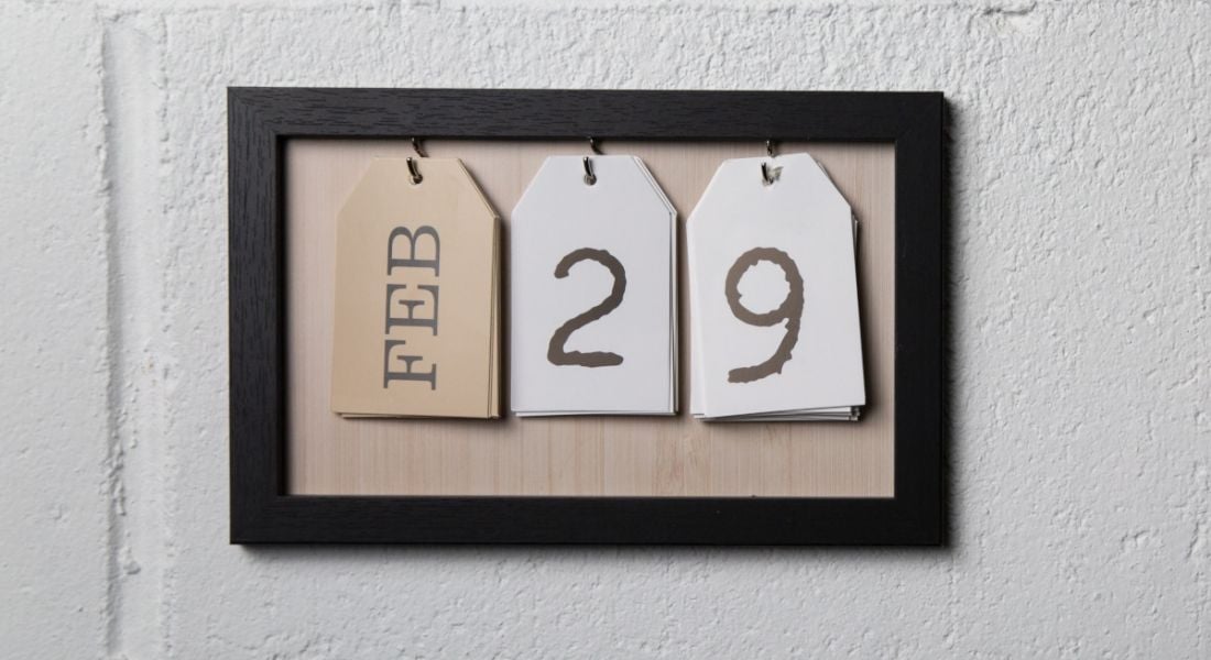 A picture frame with three tags hanging on it sits on a white wall. The first tag has the word February on it, while the other two tags spell out the number 29.