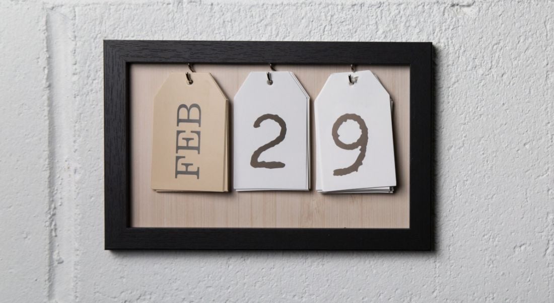 A picture frame with three tags hanging on it sits on a white wall. The first tag has the word February on it, while the other two tags spell out the number 29.