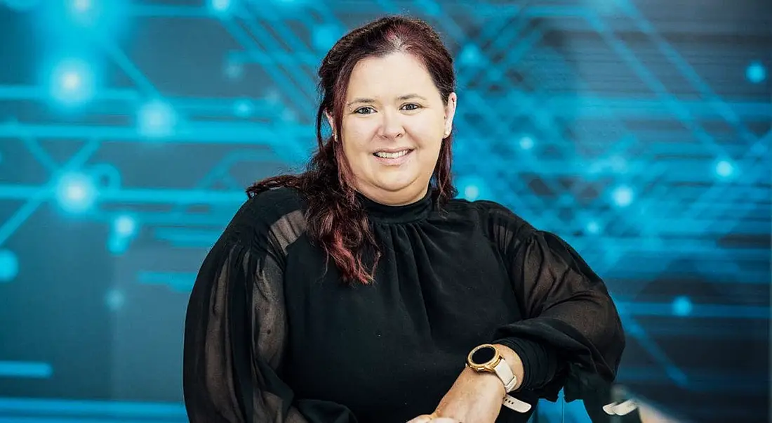 A woman smiles at the camera while resting her left elbow on a counter. There is a digital graphic behind her. She is Tricia Cahill, a cloud software engineer at Intel.