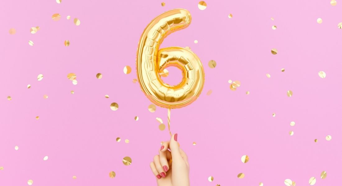 A hand with pink painted nails holding a gold balloon in the shape of a six on a pink background with sparkles.