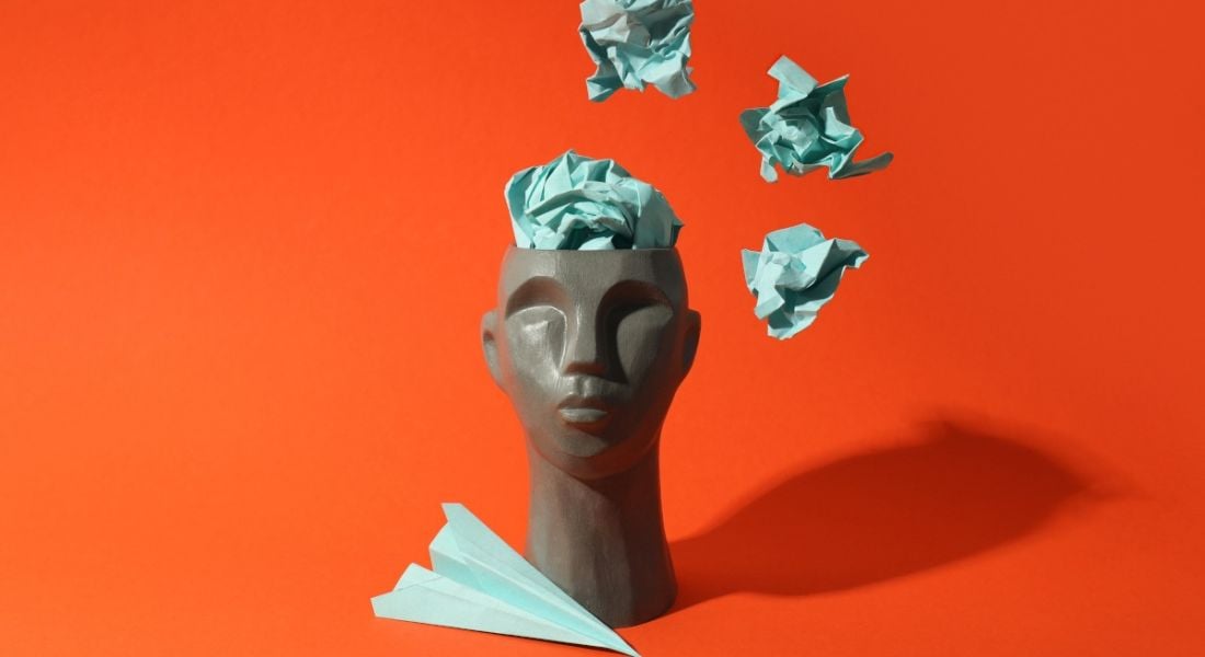 A grey head statue with a green paper plane and green balled up pieces of paper coming out of its head. The background is orange.