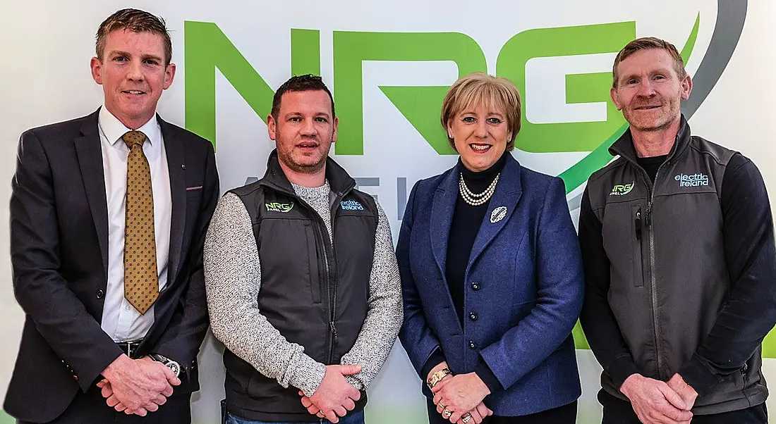 Three people from NRG Panel and Minister Heather Humphreys, TD standing in front of a sign for NRG Panel.