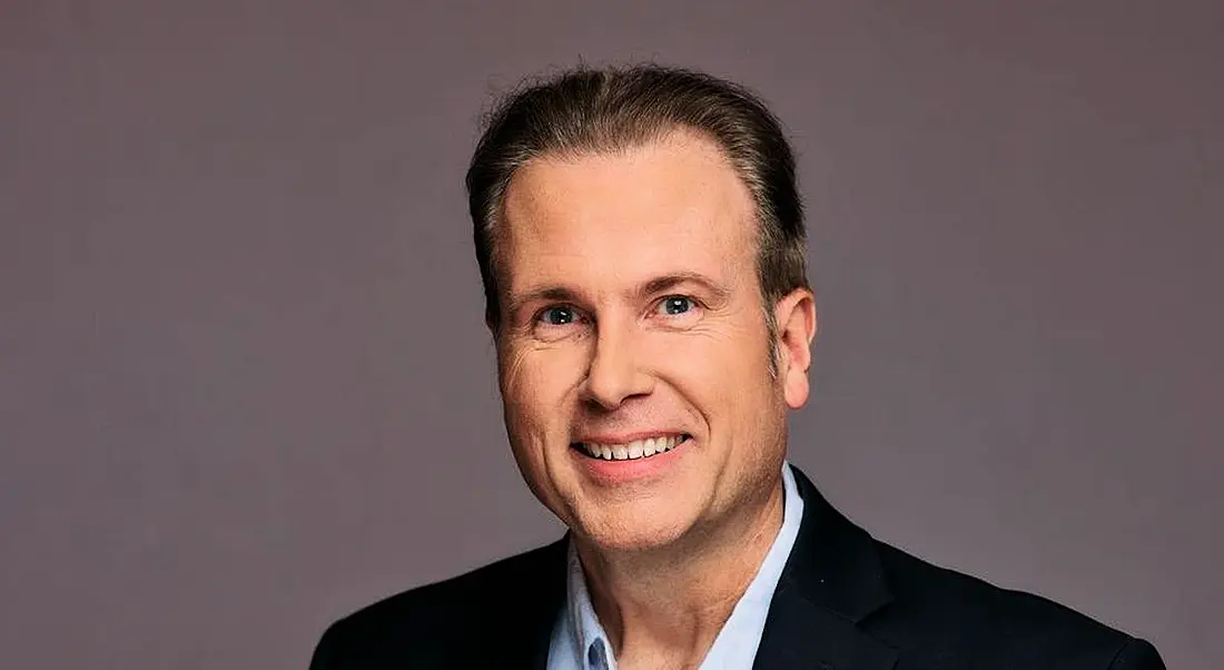 A man wearing a dark suit jacket with a white open-collared shirt smiles at the camera in front of a grey background. He is Jesse Harriott, head of analytics at Workhuman.