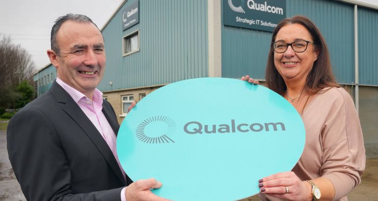 A man and a woman hold a sign that has the Qualcom logo on it. They are standing in the outdoors and in the background is a building with the Qualcom logo on it.