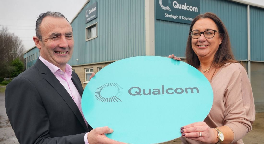 A man and a woman hold a sign that has the Qualcom logo on it. They are standing in the outdoors and in the background is a building with the Qualcom logo on it.