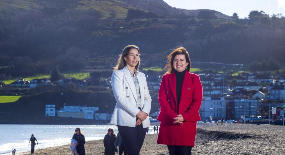 Two people from SD Worx Ireland, standing together on a beach with water and buildings of a town in the background.