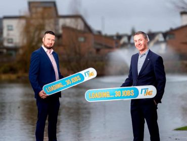 Two men wearing suits stand in front of a canal holding signs advertising 30 new jobs from IT.ie. They are Wayne Morgan and Eamon Gallagher.