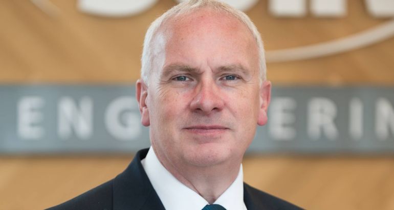 Headshot of Suir Engineering CEO John Kelly wearing a black suit and standing in front of a wall that has the Suir logo on it.