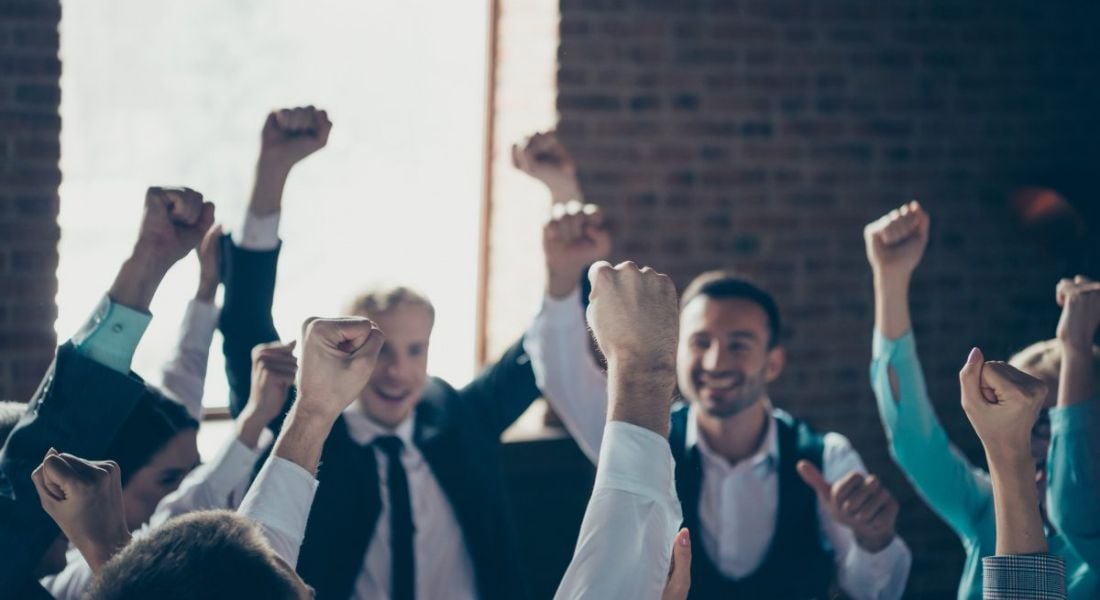 A group of employees celebrate with their arms in the air in their office.