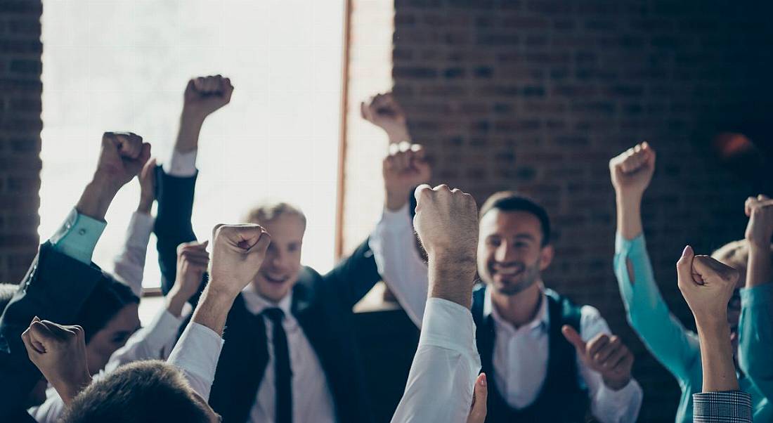 A group of employees celebrate with their arms in the air in their office.