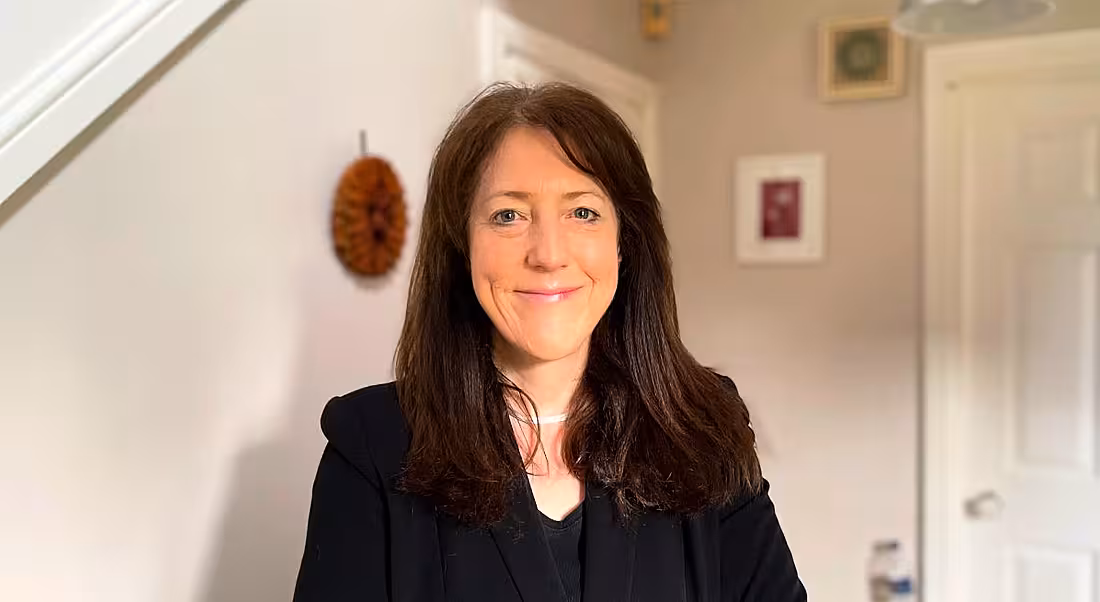 A woman with long brown hair wearing a dark blazer smiles at the camera. She is Karen MacGregor.