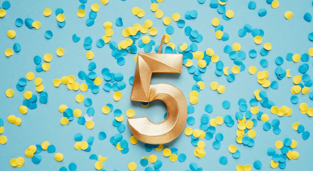 A large gold candle in the shape of the number five against a blue surface covered in yellow and blue confetti.