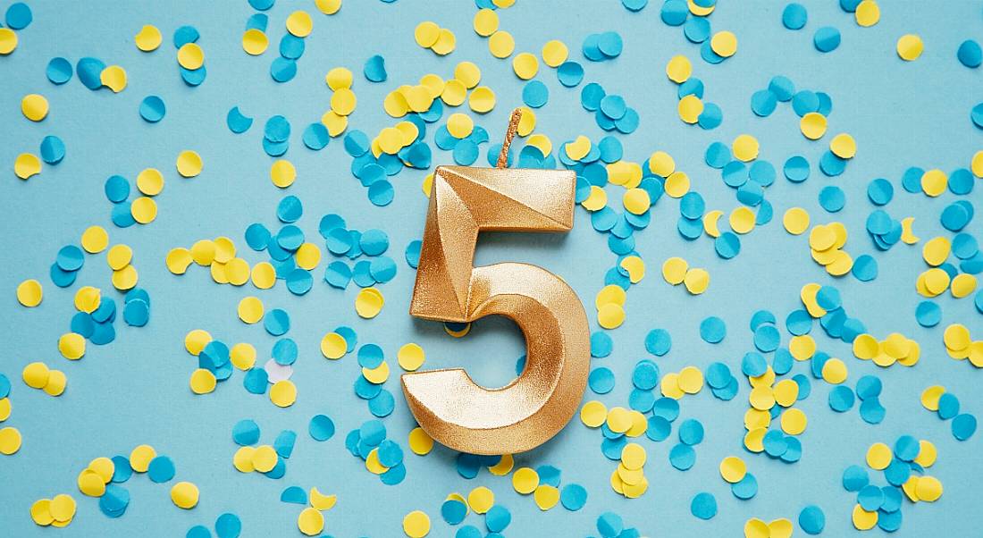 A large gold candle in the shape of the number five against a blue surface covered in yellow and blue confetti.