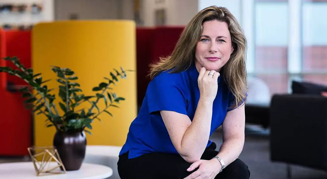 Niamh Graham sitting on the edge of a white coffee table with a plant behind her. She has her chin on her hand and is leaning forward to the camera.