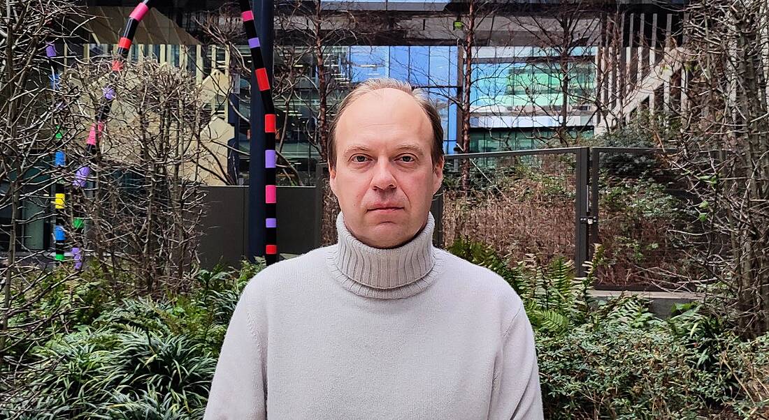 A man wearing a cream coloured turtleneck stands in a garden outside an office building. He is Oleksiy Ryabchuk, a senior software engineer at Integral Ad Science.