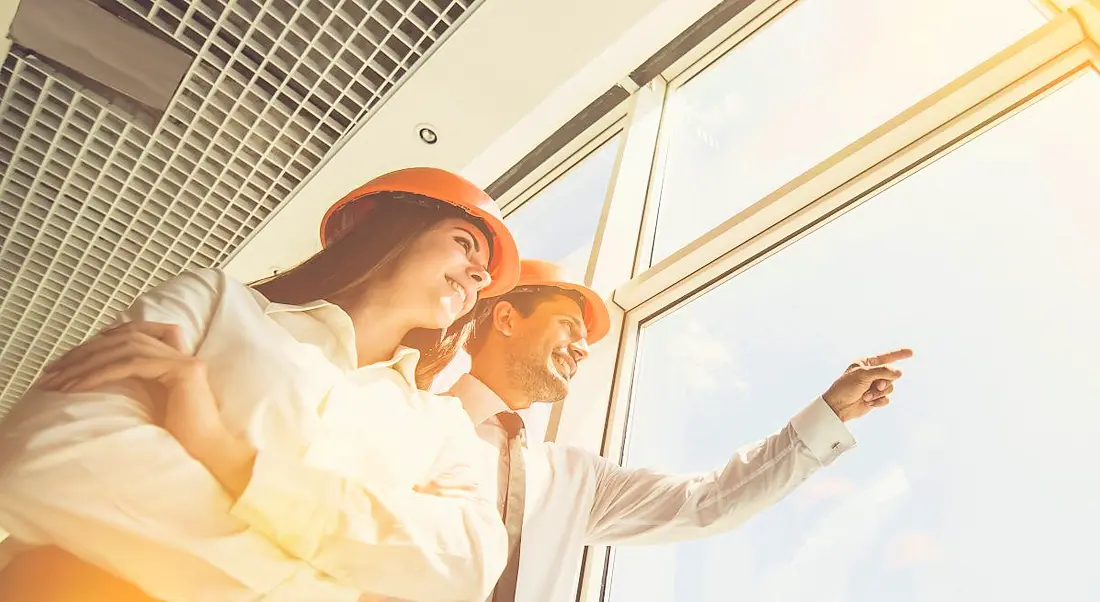 Two engineers standing beside a window with the sunlight illuminating them. One of them is pointing out the window.