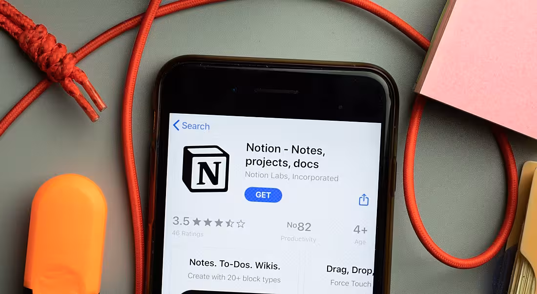 A phone displaying the Notion app download screen. It is lying on a grey table with a red cord and a pink notepad and orange eraser.