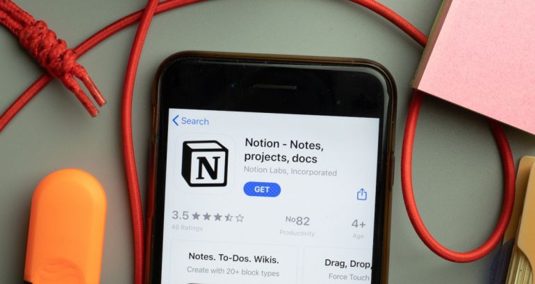 A phone displaying the Notion app download screen. It is lying on a grey table with a red cord and a pink notepad and orange eraser.