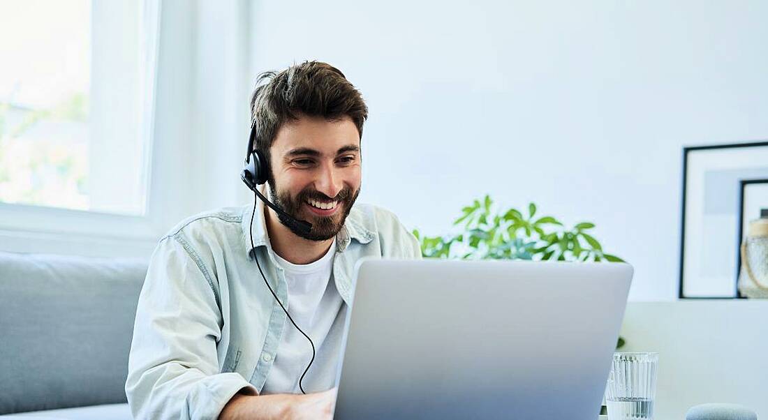 A man smiles at his laptop screen wearing a headset in a home with a couch, plants and a window behind him.